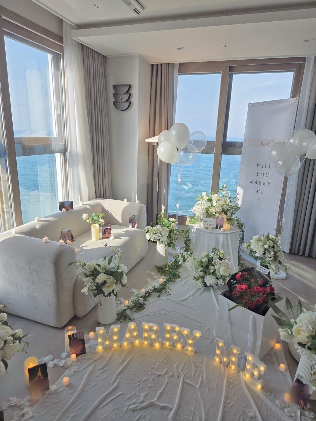 바다가 보이는 호텔 객실에 흰 꽃다발을 늘어놓고 MARRY ME 조명을 켠 프로포즈 현장